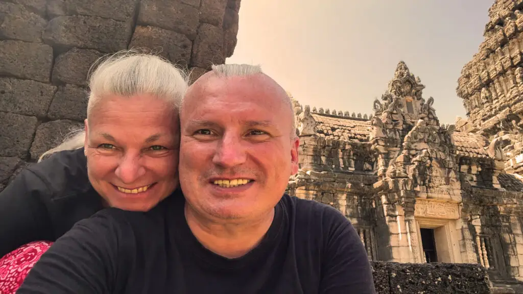Ein Selfie von Annett und Marco, den Gründern von Auswandern und Frei. Beide blicken fröhlich lächelnd in die Kamera. Sie stehen dicht beieinander vor den Ruinen eines beeindruckenden, antiken Steintempels mit aufwendigen Verzierungen und Türmen im historischen Khmer-Baustil, der in warmes Sonnenlicht getaucht ist.