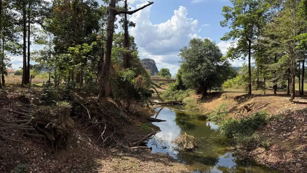 Eine authentische Landschaftsaufnahme aus Laos. Im Vordergrund fließt ein schmaler, flacher Fluss durch ein naturbelassenes Bett mit steilen Erdufern, freiliegenden Baumwurzeln und umgestürzten Ästen. Beide Ufer sind von grünen Bäumen gesäumt. Im Hintergrund führt eine Brücke über den Fluss, hinter der sich ein markanter, steiler Karstberg unter einem blauen Himmel mit weißen Schönwetterwolken erhebt.
