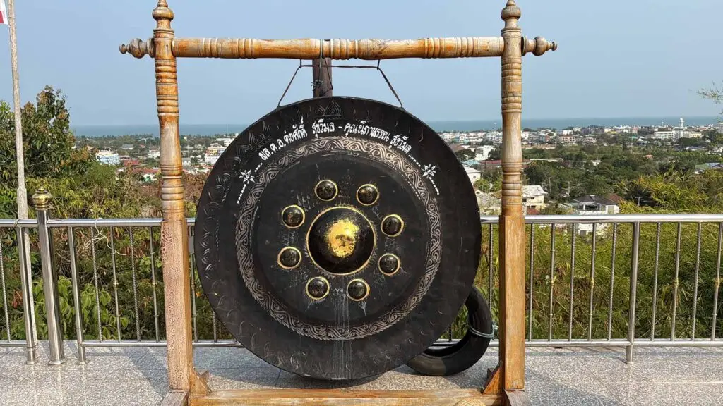 Ein großer, schwarzer thailändischer Gong mit goldenen Verzierungen und thailändischen Schriftzeichen, der an einem massiven Holzgestell hängt. Der Gong steht auf einer gefliesten Aussichtsplattform mit einem Metallgeländer. Im Hintergrund bietet sich ein weiter Panoramablick über eine thailändische Küstenstadt, grüne Landschaft und das blaue Meer unter klarem Himmel.