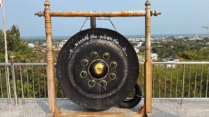 Ein großer, schwarzer thailändischer Gong mit goldenen Verzierungen und thailändischen Schriftzeichen, der an einem massiven Holzgestell hängt. Der Gong steht auf einer gefliesten Aussichtsplattform mit einem Metallgeländer. Im Hintergrund bietet sich ein weiter Panoramablick über eine thailändische Küstenstadt, grüne Landschaft und das blaue Meer unter klarem Himmel.
