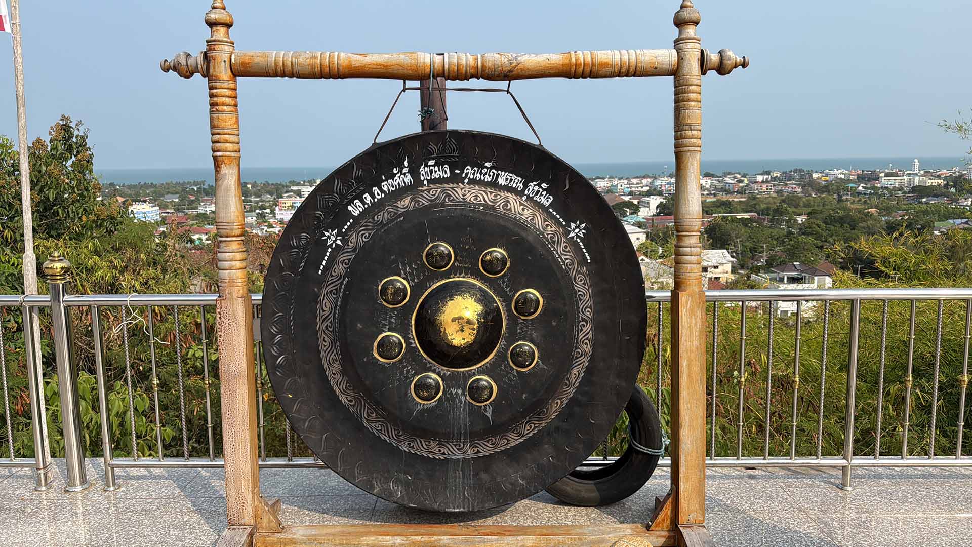 Ein großer, schwarzer thailändischer Gong mit goldenen Verzierungen und thailändischen Schriftzeichen, der an einem massiven Holzgestell hängt. Der Gong steht auf einer gefliesten Aussichtsplattform mit einem Metallgeländer. Im Hintergrund bietet sich ein weiter Panoramablick über eine thailändische Küstenstadt, grüne Landschaft und das blaue Meer unter klarem Himmel.